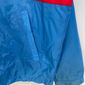 K&ouml;nnte beinhalten: Blaue Jacke mit rotem Streifen &uuml;ber der Brust und blauer Tasche. Die Jacke ist aus gl&auml;nzendem Material gefertigt.