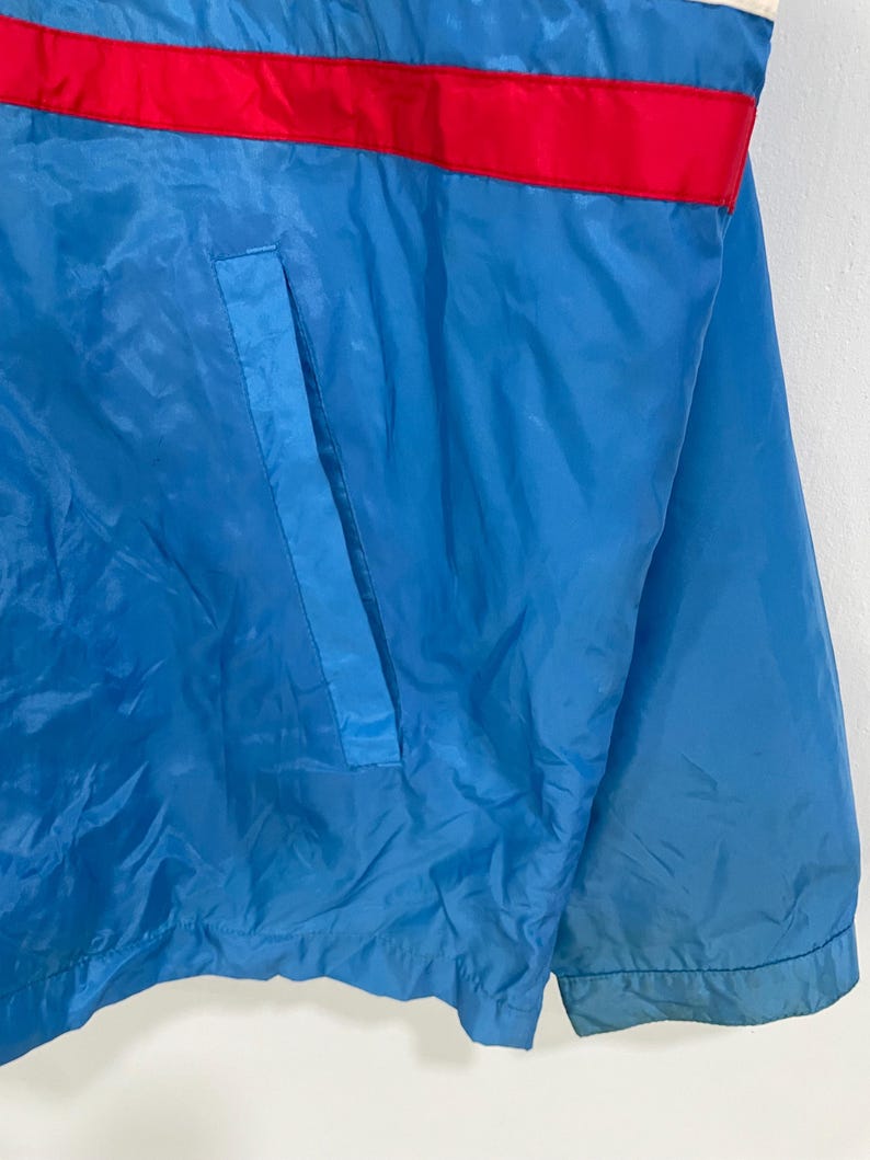 K&ouml;nnte beinhalten: Blaue Jacke mit rotem Streifen &uuml;ber der Brust und blauer Tasche. Die Jacke ist aus gl&auml;nzendem Material gefertigt.