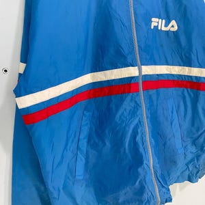 K&ouml;nnte beinhalten: Blaue FILA-Jacke mit wei&szlig;-rotem Streifendesign &uuml;ber der Brust. Die Jacke hat einen Rei&szlig;verschluss und ein kleines FILA-Logo auf der Brust. Sie hat einen Kragen und zwei Taschen.