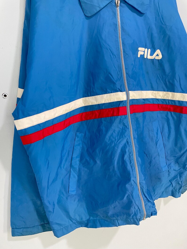 K&ouml;nnte beinhalten: Blaue FILA-Jacke mit wei&szlig;-rotem Streifendesign &uuml;ber der Brust. Die Jacke hat einen Rei&szlig;verschluss und ein kleines FILA-Logo auf der Brust. Sie hat einen Kragen und zwei Taschen.