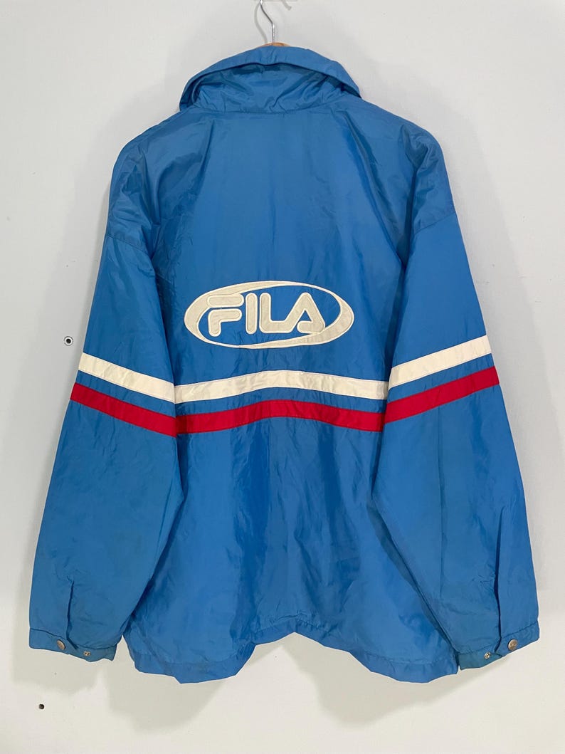 K&ouml;nnte beinhalten: Blaue Fila-Jacke mit wei&szlig;-rotem Streifendesign auf dem R&uuml;cken. Das Fila-Logo ist wei&szlig; dargestellt. Die Jacke hat einen Kragen und Knopfverschl&uuml;sse.