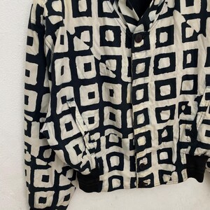 Może przedstawiać: Kurtka w stylu vintage z czarno-kremowym geometrycznym wzorem. Kurtka ma zapięcie na guziki, czarne mankiety i kołnierz. Wz&oacute;r składa się z czarnych kwadrat&oacute;w na kremowym tle.