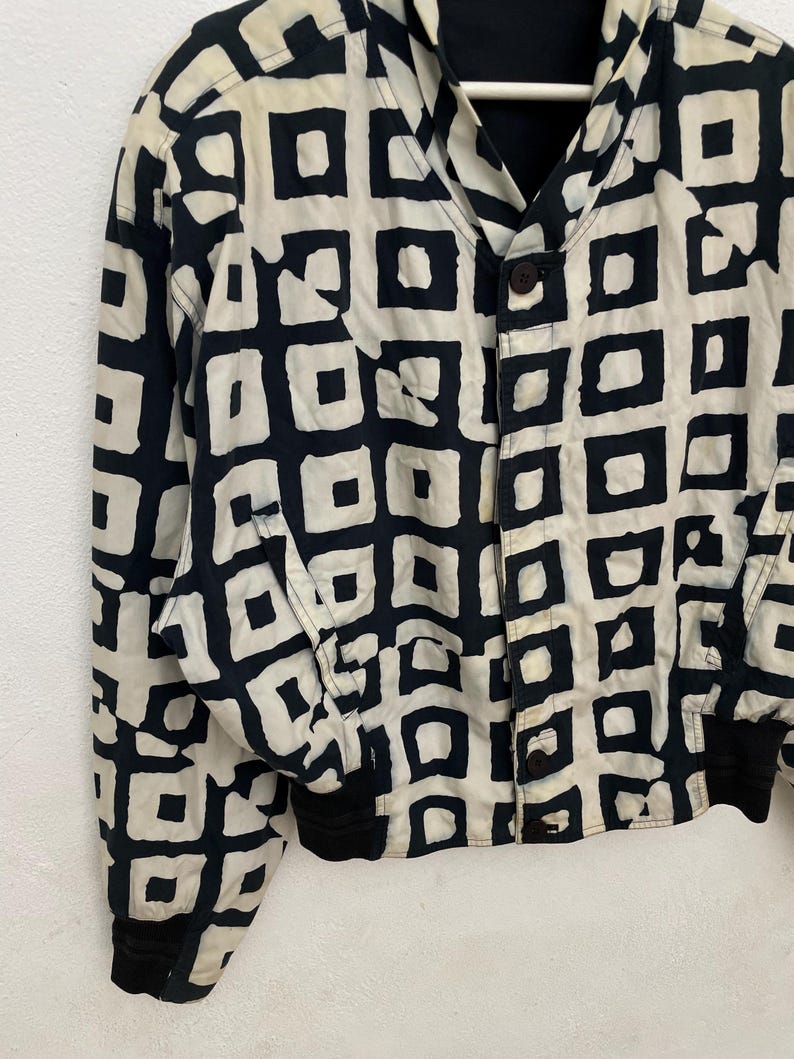 Może przedstawiać: Kurtka w stylu vintage z czarno-kremowym geometrycznym wzorem. Kurtka ma zapięcie na guziki, czarne mankiety i kołnierz. Wz&oacute;r składa się z czarnych kwadrat&oacute;w na kremowym tle.