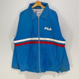 K&ouml;nnte beinhalten: Blaue Vintage-Fila-Jacke mit wei&szlig;-rotem Streifen &uuml;ber der Brust. Die Jacke hat einen durchgehenden Rei&szlig;verschluss, einen Kragen und das Fila-Logo in Wei&szlig; auf der Brust. Die Jacke h&auml;ngt an einem Kleiderb&uuml;gel.