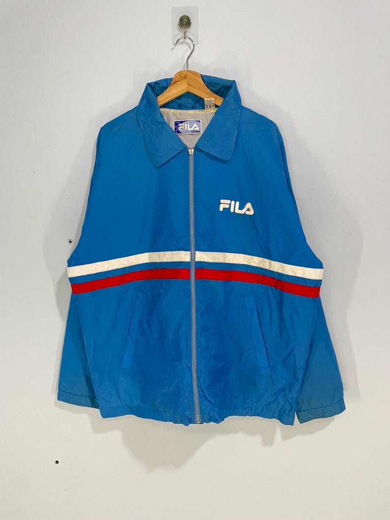 K&ouml;nnte beinhalten: Blaue Vintage-Fila-Jacke mit wei&szlig;-rotem Streifen &uuml;ber der Brust. Die Jacke hat einen durchgehenden Rei&szlig;verschluss, einen Kragen und das Fila-Logo in Wei&szlig; auf der Brust. Die Jacke h&auml;ngt an einem Kleiderb&uuml;gel.