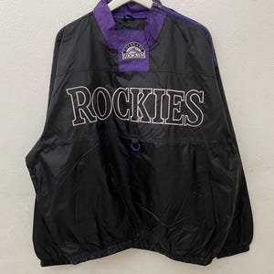 May include: Black and purple Rockies anorak jacket with the word "ROCKIES" in white lettering across the front. The jacket has a purple collar and a small logo patch. A zipper runs down the left side of the collar.