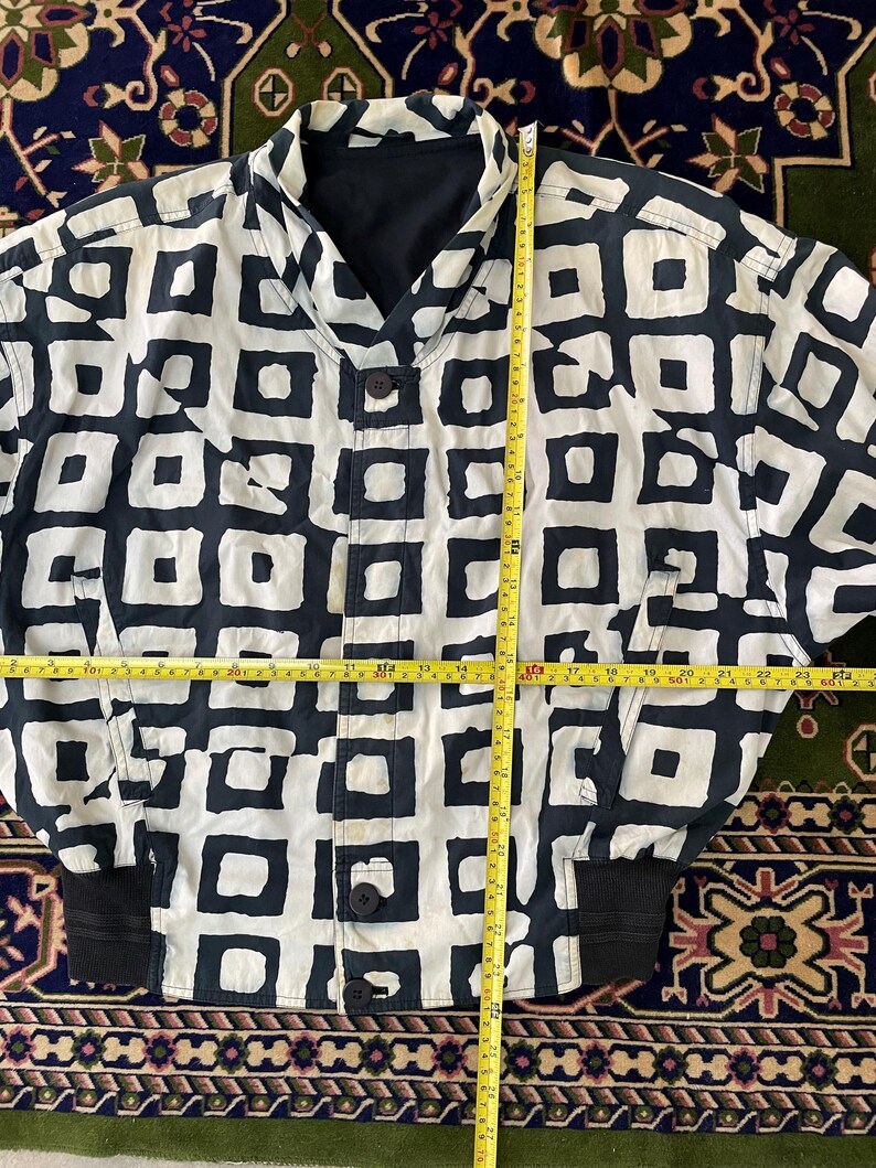 Może przedstawiać: Kurtka w stylu vintage z czarno-białym geometrycznym wzorem. Kurtka ma zapięcie na guziki, czarny kołnierz i czarne prążkowane mankiety. Kurtka ma około 71 cm długości.
