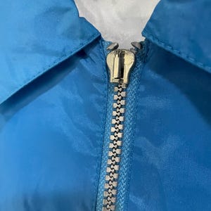 K&ouml;nnte beinhalten: Nahaufnahme einer hellblauen Jacke mit silbernem Rei&szlig;verschluss. Die Jacke hat einen Kragen und einen silbernen Rei&szlig;verschluss. Der Stoff scheint ein glattes, synthetisches Material zu sein. Die Oberseite der Jacke hat ein wei&szlig;es und blaues Etikett.
