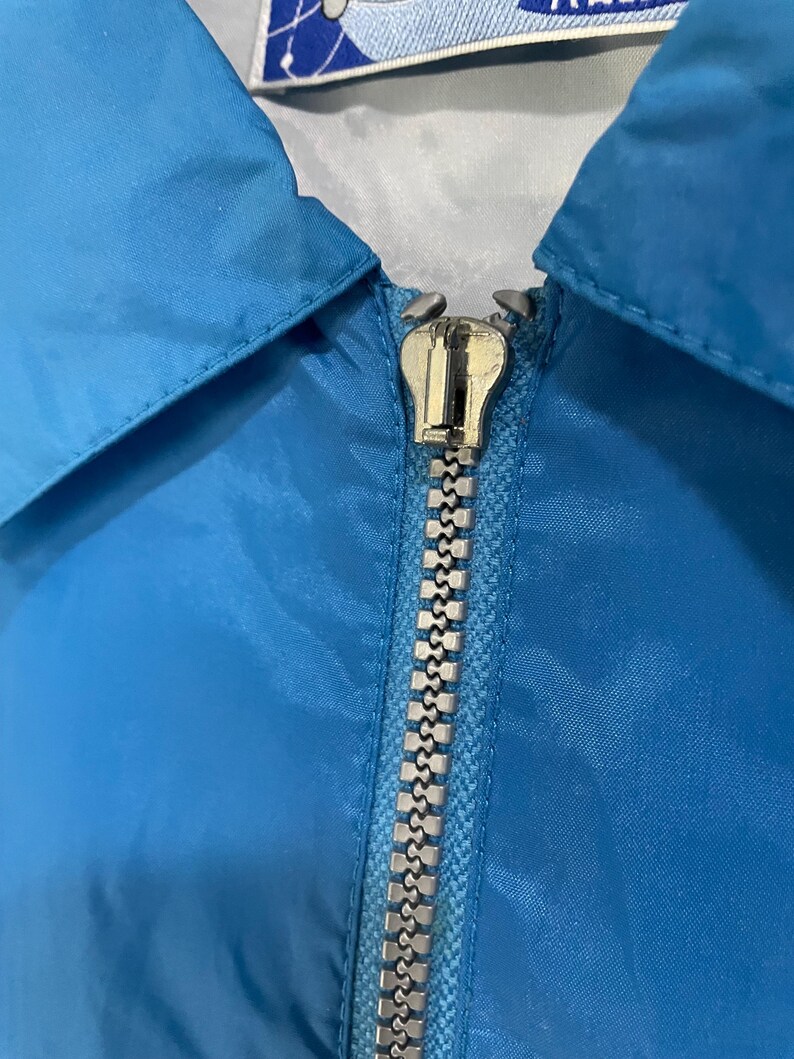 K&ouml;nnte beinhalten: Nahaufnahme einer hellblauen Jacke mit silbernem Rei&szlig;verschluss. Die Jacke hat einen Kragen und einen silbernen Rei&szlig;verschluss. Der Stoff scheint ein glattes, synthetisches Material zu sein. Die Oberseite der Jacke hat ein wei&szlig;es und blaues Etikett.