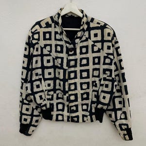 Puede incluir: Chaqueta negra y crema con un patrón geométrico de cuadrados. La chaqueta tiene una parte delantera abotonada, puños negros y un cuello negro. Cuelga de una percha blanca sobre un fondo blanco.