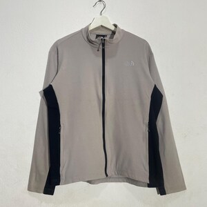May include: A light gray zip-up jacket with black side panels and a small logo on the chest. The jacket has a high collar and two zippered pockets. The North Face brand jacket is hanging on a white background.