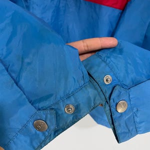 K&ouml;nnte beinhalten: Nahaufnahme einer blauen Jacke mit roten und wei&szlig;en Streifen &uuml;ber der Brust. Die Jacke hat Druckkn&ouml;pfe an den &Auml;rmeln und der Markenname ist auf den Druckkn&ouml;pfen sichtbar.