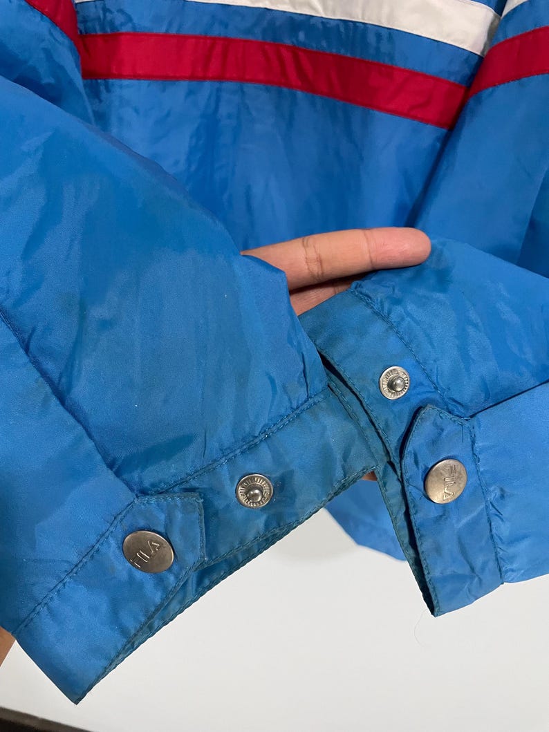 K&ouml;nnte beinhalten: Nahaufnahme einer blauen Jacke mit roten und wei&szlig;en Streifen &uuml;ber der Brust. Die Jacke hat Druckkn&ouml;pfe an den &Auml;rmeln und der Markenname ist auf den Druckkn&ouml;pfen sichtbar.