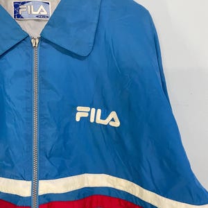 K&ouml;nnte beinhalten: Blaue Fila-Jacke mit wei&szlig;em Logo und Rei&szlig;verschluss. Die Jacke hat wei&szlig;e und rote Streifen &uuml;ber der Brust. Der Kragen ist hellblau. Auf dem Etikett steht "FILA ITALIA".