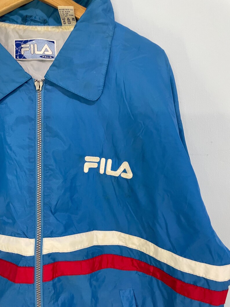 K&ouml;nnte beinhalten: Blaue Fila-Jacke mit wei&szlig;em Logo und Rei&szlig;verschluss. Die Jacke hat wei&szlig;e und rote Streifen &uuml;ber der Brust. Der Kragen ist hellblau. Auf dem Etikett steht "FILA ITALIA".
