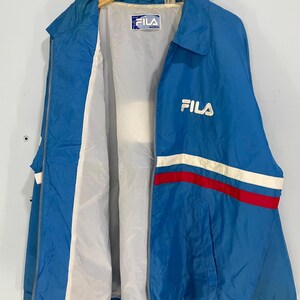 K&ouml;nnte beinhalten: Blaue Fila-Jacke mit wei&szlig;-rotem Streifen &uuml;ber der Brust. Die Jacke hat einen durchgehenden Rei&szlig;verschluss und ein wei&szlig;es Innenfutter. Das Fila-Logo ist auf der Brust und im Kragen sichtbar. Die Jacke h&auml;ngt an einem Holzb&uuml;gel.