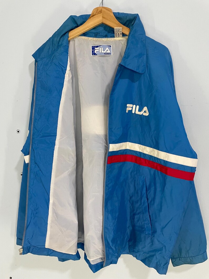 K&ouml;nnte beinhalten: Blaue Fila-Jacke mit wei&szlig;-rotem Streifen &uuml;ber der Brust. Die Jacke hat einen durchgehenden Rei&szlig;verschluss und ein wei&szlig;es Innenfutter. Das Fila-Logo ist auf der Brust und im Kragen sichtbar. Die Jacke h&auml;ngt an einem Holzb&uuml;gel.