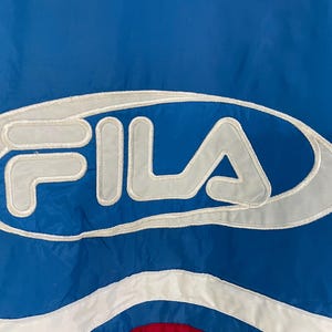 K&ouml;nnte beinhalten: Nahaufnahme einer blau-wei&szlig;en Fila-Jacke. Das Fila-Logo ist wei&szlig; mit grauem Umriss dargestellt. Die Jacke hat einen blauen Hintergrund mit wei&szlig;en und roten Akzenten.