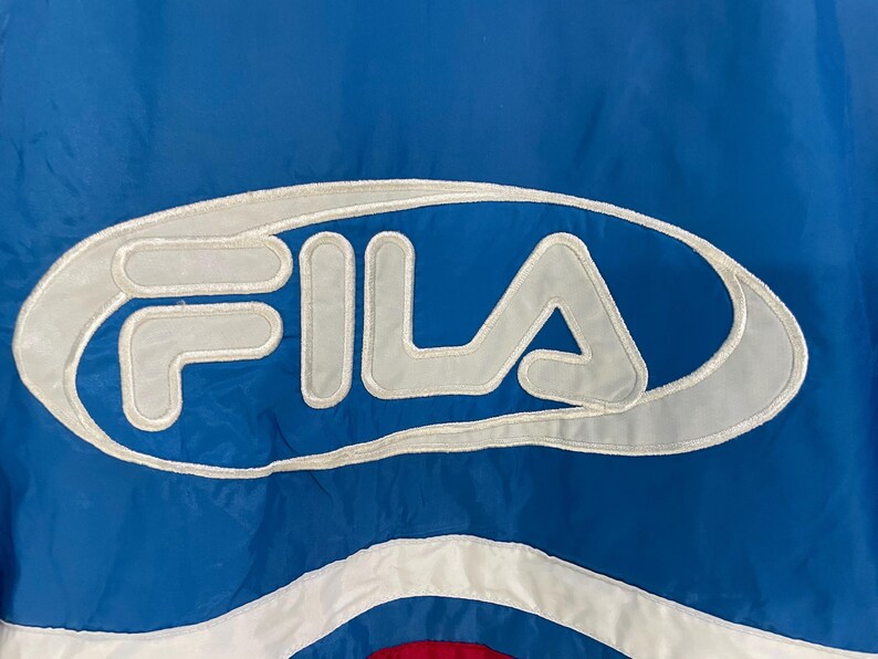 K&ouml;nnte beinhalten: Nahaufnahme einer blau-wei&szlig;en Fila-Jacke. Das Fila-Logo ist wei&szlig; mit grauem Umriss dargestellt. Die Jacke hat einen blauen Hintergrund mit wei&szlig;en und roten Akzenten.