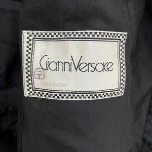 Może przedstawiać: Zbliżenie czarnego ubrania z etykietą. Etykieta zawiera napis "Gianni Versace" w eleganckiej czcionce, obramowany szachownicą. Etykieta zawiera r&oacute;wnież napis "MADE IN ITALY".