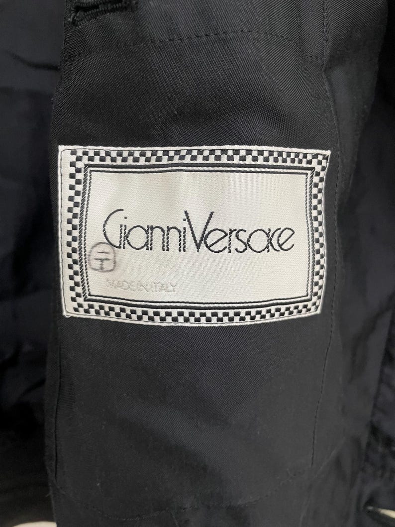 Może przedstawiać: Zbliżenie czarnego ubrania z etykietą. Etykieta zawiera napis "Gianni Versace" w eleganckiej czcionce, obramowany szachownicą. Etykieta zawiera r&oacute;wnież napis "MADE IN ITALY".