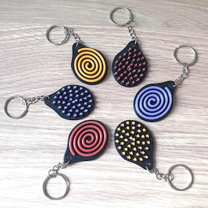 May include: Six keyrings with black teardrop-shaped pendants. Each keyring has a different design: a yellow spiral, red dots, blue dots, a purple spiral, a red spiral, and yellow dots. The keyrings have silver-coloured key rings.