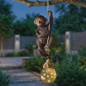 May include: A brown monkey ornament suspended from a tree branch by a rope. The monkey figure is holding the rope. A glass globe with small lights hangs below the monkey, creating a decorative outdoor light feature.
