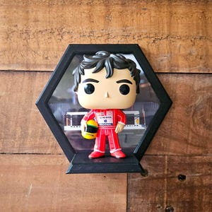 Op de afbeelding: Een Funko Pop-figuur van een autocoureur in een rood racepak, met een gele en zwarte helm, tentoongesteld in een zwarte zeshoekige schaduwbox. De achtergrond toont een wazige afbeelding van een racebaan. De schaduwbox is op een houten oppervlak gemonteerd.