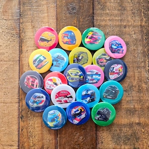 May include: A selection of circular badges displaying racing helmet designs in a variety of colours. The badges, in shades like red, yellow, blue, green, and pink, are arranged on a wooden surface.