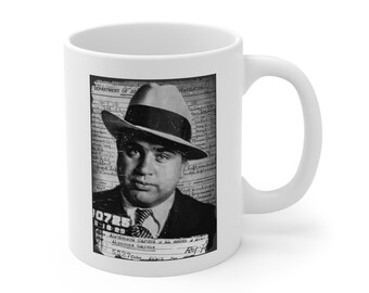 May include: White ceramic mug featuring a black and white image of a vintage mugshot of a man in a fedora. The mugshot is set against a background of text, with the name "Alphonse Capone" visible.