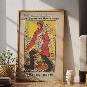 Può includere: Un poster incorniciato in stile vintage raffigurante una figura centrale con un cappotto rosso che trasporta una grande pila di fogli su uno sfondo giallo. Il testo recita "День Советской Пропаганды" in alto e "ЗНАНИЕ - ВСЕМ!" in basso, con una scena urbana.