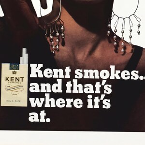 May include: Vintage advertisement featuring a pack of Kent cigarettes, with the text "Kent smokes... and that's where it's at." The image includes a woman wearing earrings. The cigarette pack is beige with the brand name in black.