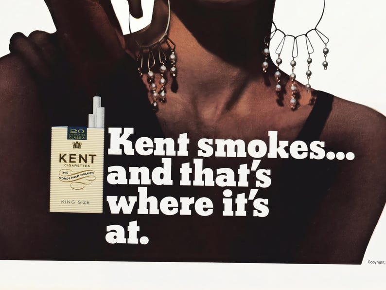 May include: Vintage advertisement featuring a pack of Kent cigarettes, with the text "Kent smokes... and that's where it's at." The image includes a woman wearing earrings. The cigarette pack is beige with the brand name in black.