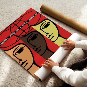 May include: A rectangular poster featuring a stylized graphic illustration of three faces in shades of yellow, brown, and tan against a vibrant red background. The text "MARZO 8" and "MARCH 8th" is printed in black across the upper section.
