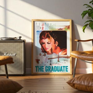 May include: A framed movie poster for The Graduate featuring a close-up illustration of two people, a depiction of the Golden Gate Bridge, and Japanese text. The poster is displayed in a light wood frame with a white border and bold blue text.