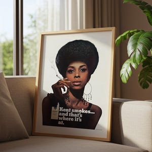 May include: Framed vintage advertisement featuring a woman with a large afro smoking a cigarette. The poster has the text "Kent smokes... and that's where it's at." The image is in a wooden frame, resting on a sofa.