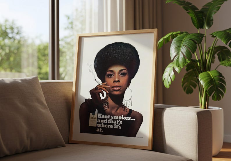 May include: Framed vintage advertisement featuring a woman with a large afro smoking a cigarette. The poster has the text "Kent smokes... and that's where it's at." The image is in a wooden frame, resting on a sofa.