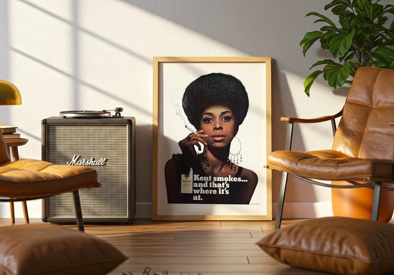 May include: A framed vintage poster featuring a woman with a large afro smoking a cigarette. The poster has the text "Kent smokes... and that's where it's at." A Marshall speaker and a record player are next to the poster. A leather chair and a plant are also in the room.