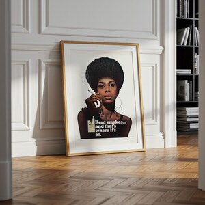 May include: Framed vintage advertisement featuring a woman with a large afro smoking a cigarette. The print has a gold frame and rests against a white wall. The text on the print reads "Kent smokes... and that's where it's at."