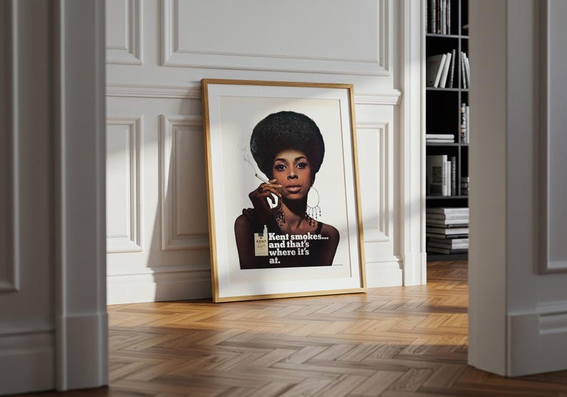 May include: Framed vintage advertisement featuring a woman with a large afro smoking a cigarette. The print has a gold frame and rests against a white wall. The text on the print reads "Kent smokes... and that's where it's at."