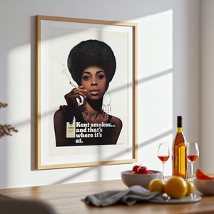 May include: Framed vintage advertisement featuring a woman with a large afro smoking a cigarette. The ad reads "Kent smokes... and that's where it's at." A bottle of wine, glasses, and fruit bowls are on a wooden table.