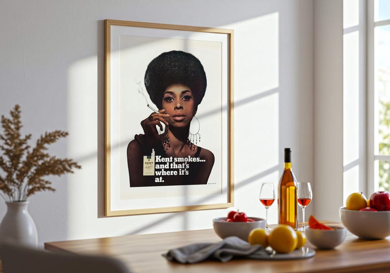 May include: Framed vintage advertisement featuring a woman with a large afro smoking a cigarette. The ad reads "Kent smokes... and that's where it's at." A bottle of wine, glasses, and fruit bowls are on a wooden table.