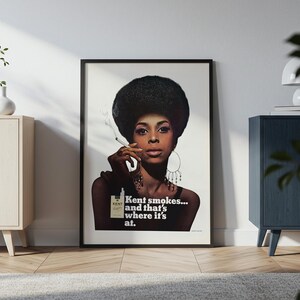 May include: Framed vintage advertisement featuring a woman with a large afro holding a cigarette. The ad for Kent cigarettes includes the text "Kent smokes... and that's where it's at." The image is in a black frame, leaning against a white wall.