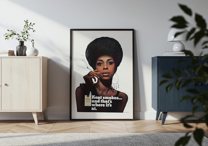 May include: Framed vintage advertisement featuring a woman with a large afro holding a cigarette. The ad for Kent cigarettes includes the text "Kent smokes... and that's where it's at." The image is in a black frame, leaning against a white wall.