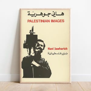 May include: A framed poster with a beige background features a black and red graphic. The graphic depicts a man holding a camera, with text in Arabic and English that reads "PALESTINIAN IMAGES" and "Hani Jawharieh."