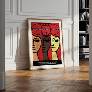 May include: A framed poster featuring a stylized graphic illustration of three faces in tan, brown, and yellow against a red background. Above the faces is a globe outline and the text "MARZO 8 MARCH 8th". Below is "DIA INTERNACIONAL DE LA MUJER INTERNATIONAL WOMEN'S DAY".