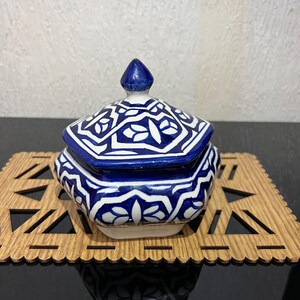 Peut inclure: Un pot en céramique bleu et blanc avec un couvercle et un motif géométrique. Le pot a un bouton pointu et repose sur un dessous de plat en bois avec un bord décoratif. C'est un objet décoratif.