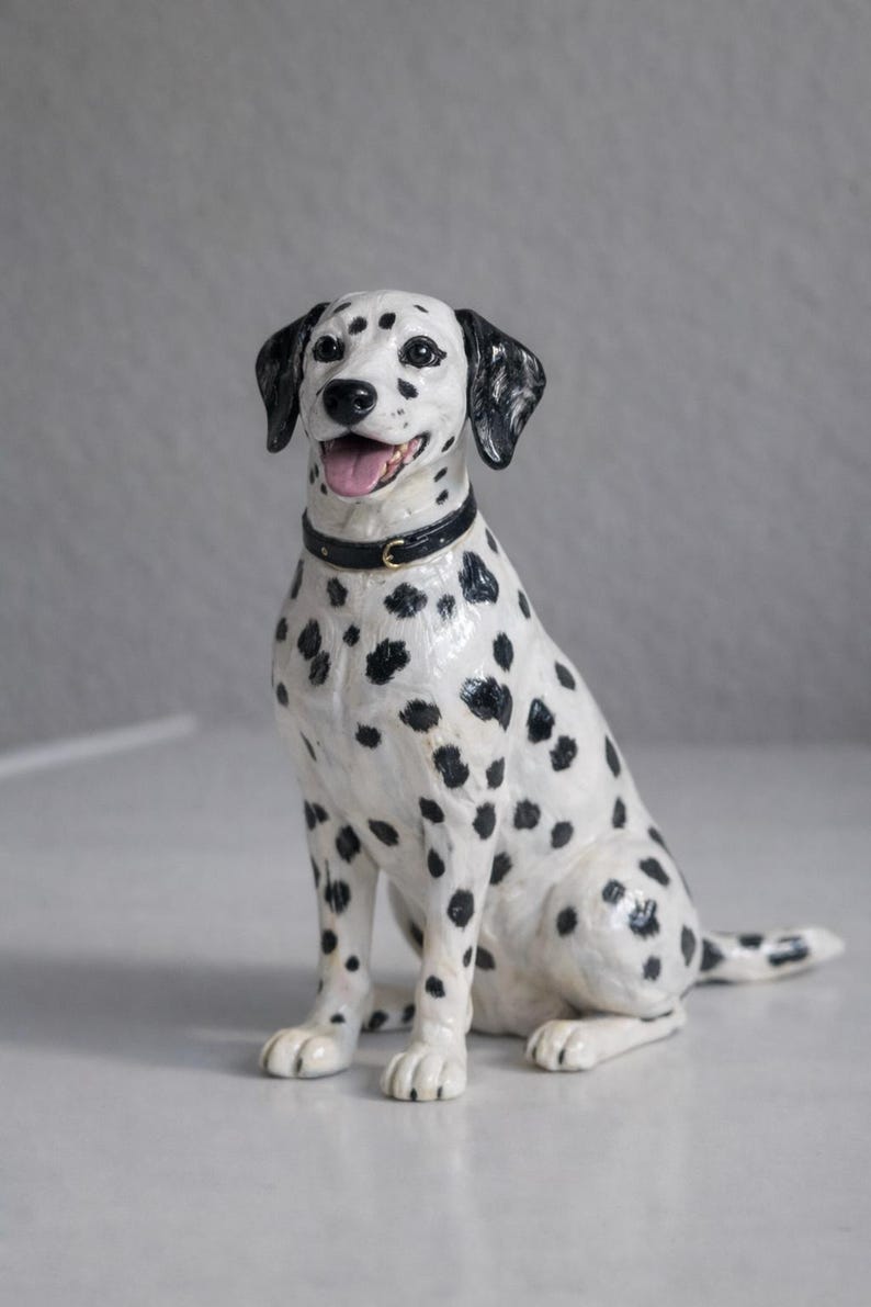 Może przedstawiać: Ceramiczna figurka psa dalmatyńczyka, siedzącego z wystawionym językiem. Pies jest biały z czarnymi plamami i ma czarną obrożę. Figurka to przedmiot dekoracyjny.