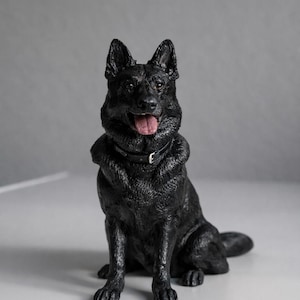 Może przedstawiać: Figurka czarnego owczarka niemieckiego, siedzącego z wystawionym językiem. Pies ma obrożę z klamrą. Figurka wykonana jest z teksturowanego materiału i osadzona na szarym tle.