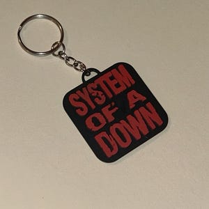 May include: A black square keychain with the words "SYSTEM OF A DOWN" in red block letters. The keychain has a silver metal ring and chain. The background is a light, neutral color.